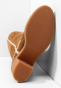 Botte en daim marron avec semelle en caoutchouc épaisse inclinée contre un mur blanc, montrant le dessous texturé et les coutures détaillées sur la partie supérieure.