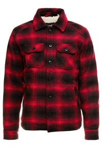Veste en flanelle à carreaux rouges et noirs avec un devant boutonné, deux poches poitrine et un col. Texture douce et doublure chaude.