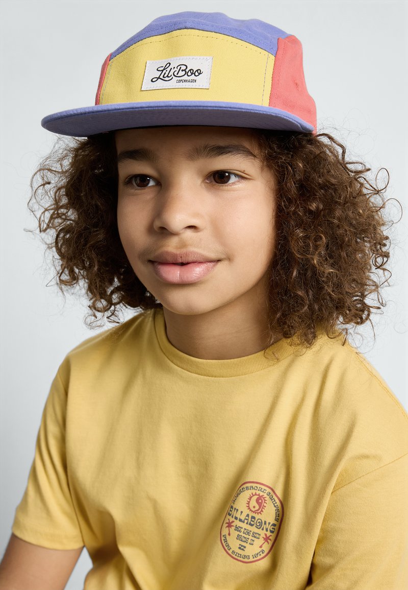 Bunte Kappe mit lila, gelben und roten Panels; verfügt über ein Marken-Patch. Das Kind trägt ein schlichtes gelbes T-Shirt mit einem Logo.