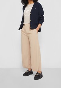 Marinblå stickad kavaj med guldknappar, beige grafisk T-shirt, vida beige byxor och svarta loafers med guldfärgade detaljer.