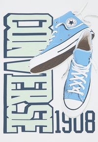 Blå høye sneakers med hvite snorer og gummisåler plassert over et grafisk design med "CONVERSE" og "1908" i fet skrift.