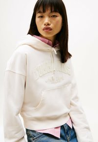 Felpa beige chiaro con tasca a marsupio e logo ricamato "Tommy Jeans New York". Indossata sopra una camicia rosa con colletto, accompagnata da jeans in denim.