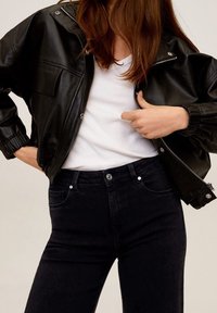 Schwarze Jacke aus Kunstleder mit Reißverschluss und elastischen Bündchen, getragen über einem weißen Hemd, kombiniert mit dunklen Jeans.