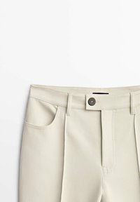 Pantalon beige avec une texture lisse, doté d'une fermeture à bouton, de passants de ceinture et de poches latérales. Le design comprend un pli sur le devant.