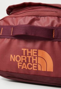 Sac bordeaux avec une finition texturée, arborant un logo et un graphisme orange. Il possède une sangle renforcée et une fermeture zippée sur le dessus.