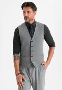 Mann in grauem Weste und Hose mit schwarzem Hemd und Krawatte, steht mit einer Hand in der Tasche, Tätowierungen sind auf den Unterarmen sichtbar.