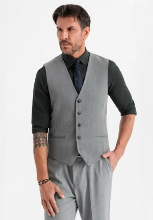 Uomo in panciotto e pantaloni grigi con camicia nera e cravatta, in piedi con una mano in tasca, tatuaggi visibili sugli avambracci.