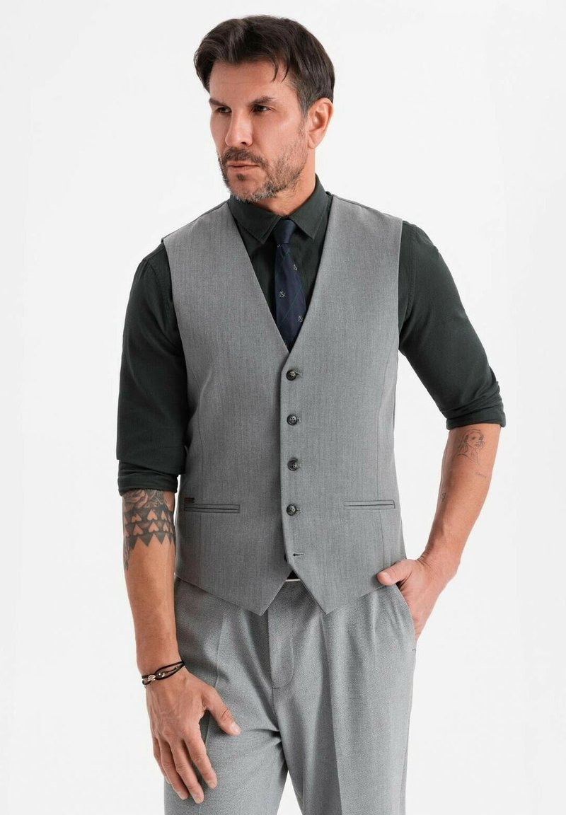 Mann in grauem Weste und Hose mit schwarzem Hemd und Krawatte, steht mit einer Hand in der Tasche, Tätowierungen sind auf den Unterarmen sichtbar.