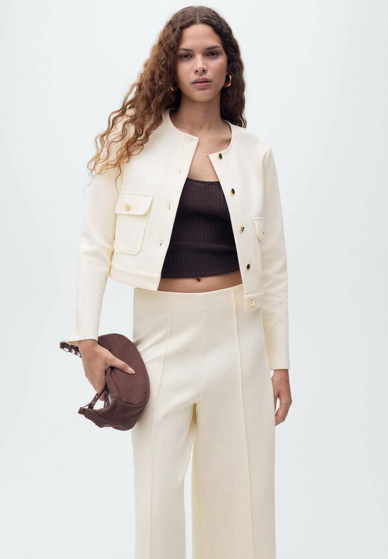 Giacca bianca corta con bottoni dorati, abbinata a un top marrone a coste e pantaloni crema. La modella tiene una borsa marrone, che mostra un design pulito.