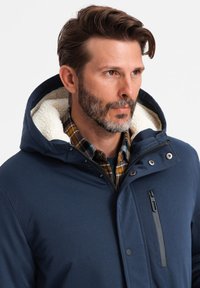 Parka blu navy con cappuccio foderato in pelliccia sintetica, chiusura con zip e bottoni a pressione, e una camicia a quadri sotto. Presenta una tasca sul petto con zip.