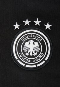 Czarny materiał z okrągłą haftowaną naszywką z białym emblematem orła, otoczoną czterema białymi gwiazdami i napisem "Deutscher Fussball-Bund."