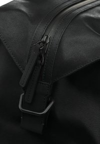 Sac à dos noir montrant un tissu texturé, une fermeture éclair lisse et des accents en cuir. Les caractéristiques incluent une boucle en métal et des coutures détaillées.