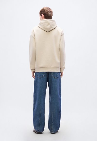 Sweat à capuche beige associé à un pantalon en denim bleu à jambes larges, présentant un design décontracté et des poches arrière visibles.