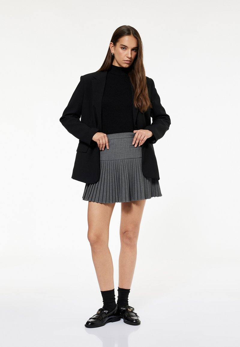 Grå plisseret mini nederdel med en flad talje, parret med en sort turtleneck og en sort blazer; afsluttet med glinsende sorte loafers.