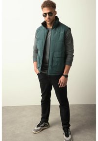 Grüne Pufferweste mit hohem Kragen, schwarzem Innenfutter, zwei Fronttaschen, getragen über einem grauen Langarmshirt, kombiniert mit schwarzen Jeans und Sneakers.