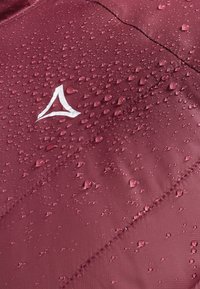 Bordeauxfarbene wasserdichte Jacke aus Textil mit strukturierter Oberfläche, versehen mit Wassertropfen und einem weißen Logo in der oberen linken Ecke.