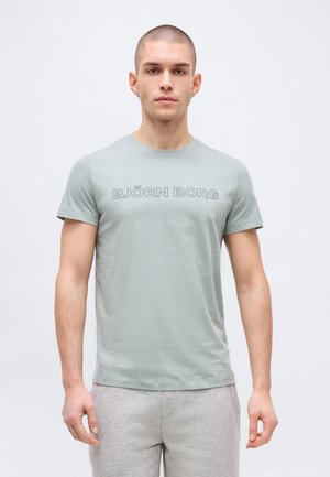 Vyras trumpais plaukais, dėvi šviesiai žalią Björn Borg marškinėlį ir šviesiai pilkas sportines kelnes, stovi priešais paprastą baltą foną.