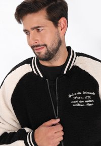 Sorte og hvide fleece bomberjakke med ribbede manchetter og halsudskæring; har lynlås lukning og broderet tekst på forsiden.