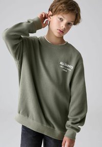 Pullover in maglia a coste verde con scollatura a girocollo, maniche lunghe e polsini a costina. Presenta un logo bianco stampato sul petto. Abbinato a un jeans scuro.