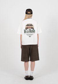 Person som har på seg en hvit oversized T-skjorte med en vintagebil og teksten "In a Rush International" på baksiden, mørke shorts, hvite sokker og svart caps.