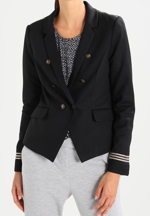 Blazer negro de corte entallado, con botones plateados, dos bolsillos delanteros y detalles a rayas en las mangas, llevado sobre una blusa estampada.