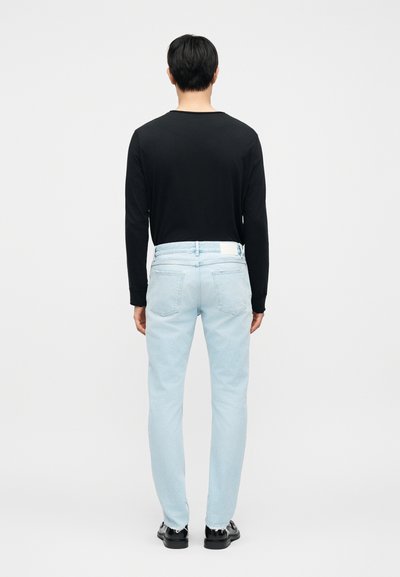 Άτομο που φοράει ανοιχτό μπλε τζιν και μαύρο μακρυμάνικο πουκάμισο, όρθιο με την πλάτη προς την κάμερα, μπροστά σε μονόχρωμο λευκό φόντο.