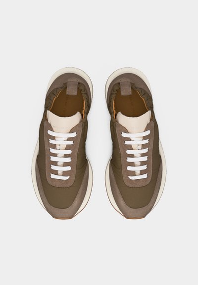 Ζευγάρι αθλητικών παπουτσιών σε ελιά και μπεζ χρώμα, με λευκά κορδόνια και χοντρές λευκές σόλες, φωτογραφημένα από πάνω πάνω σε ανοιχτό γκρι φόντο.
