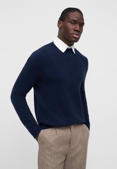 Μπλε πουλόβερ με στρογγυλή λαιμόκοψη, κατασκευασμένο από απαλό υλικό, συνδυασμένο με ανοιχτόχρωμο, υφασμάτινο παντελόνι και λευκό πουκάμισο με γιακά.