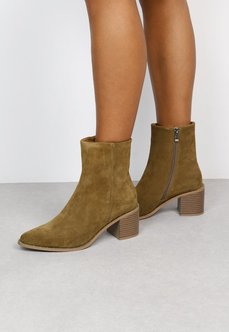 Botas de tobillo de ante marrón con punta afilada, cremallera lateral y un tacón apilado texturizado. Superficie suave con un detalle mínimo.