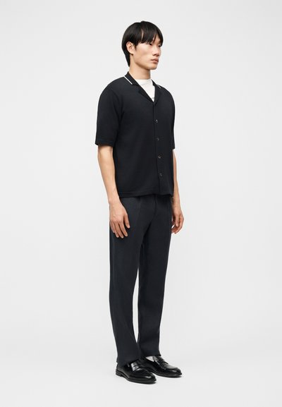 Jeune homme aux cheveux noirs courts, debout de profil à droite, portant une chemise noire à manches courtes boutonnée, un pantalon noir et des chaussures noires.