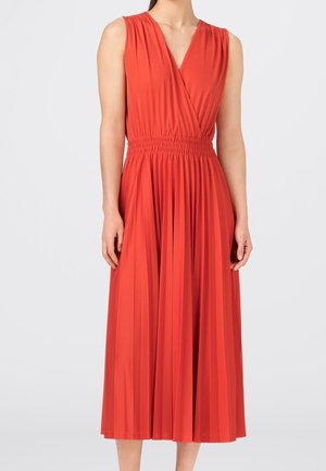 Vestido midi sin mangas de color rojo-anaranjado con escote en V, cintura fruncida y falda plisada, llevado por una persona que está de pie frente a un fondo liso.