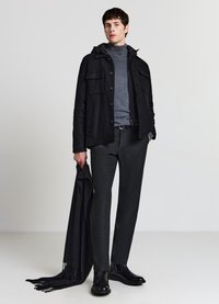 Schwarze Jacke aus Wollmischung mit Kapuze und mehreren Vordertaschen, grauer Rollkragenpullover, strukturierte dunkle Hose und schwarze Stiefeletten.