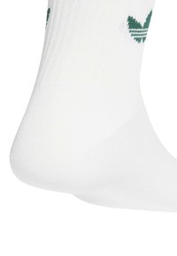 Calcetín blanco de punto hasta el tobillo con el logotipo trefoil verde de Adidas cerca del puño, tela texturizada y áreas del talón y los dedos reforzadas.