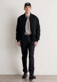 Chaqueta bomber negra sobre una camisa de botones gris claro, combinada con pantalones y zapatos negros. Una gorra negra presenta un logo en la parte frontal.