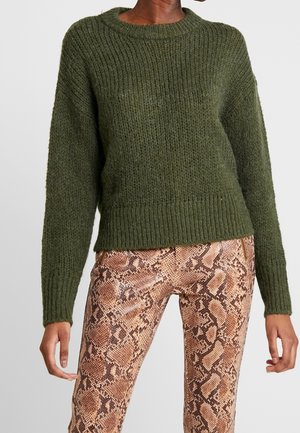 Personne portant un pull en maille épaisse vert olive et un pantalon en imprimé serpent marron, se tenant contre un fond clair.
