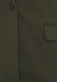 Tela de abrigo verde oscuro con un gran botón cerca del cuello y un bolsillo con solapa en el lado derecho.