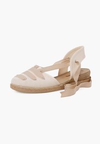 Espadrille de lienzo beige con suela de yute trenzada, cintas cruzadas sobre los dedos y lazos de cinta en el tobillo. Textura lisa con un ligero brillo.