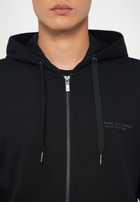 Crna zip-sweat majica s mekim teksturom, sadrži srebrni patentni zatvarač i logo detalj na prsima.Ukućane špice su od odgovarajućeg materijala.