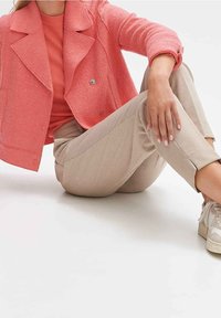 Personne assise avec un genou plié, portant une veste texturée corail, un haut assorti, un pantalon beige et des baskets blanches sur fond blanc.