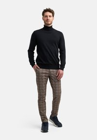 Hombre con un suéter negro y pantalones de cuadros marrones se encuentra de pie con una mano en el bolsillo, frente a un fondo blanco, sonriendo con confianza.