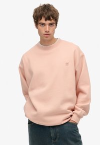Felpa rosa chiaro con scollatura a girocollo, design a spalle scese e un piccolo logo sul petto. Realizzata in tessuto morbido e texturizzato.