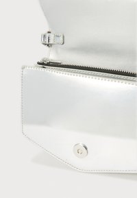 Ασημένια μεταλλική τσάντα με λεία υφή, θήκη με φερμουάρ και κλείσιμο με κόπιτσα. Διαθέτει εξαγωνικό σχήμα και αντίθετο μαύρο φερμουάρ.