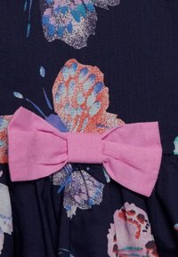 Vaaleanpunainen kangasruusu kiinnitetty tumman kukkakuosiselle kankaalle, jossa on sinisiä, purppuran värisiä ja korallin sävyisiä abstrakteja kukkamuotoja.