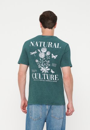 Homme portant un T-shirt vert avec des imprimés floraux et d'insectes blancs et les mots "Natural Culture" dans le dos, associé à un jean bleu.