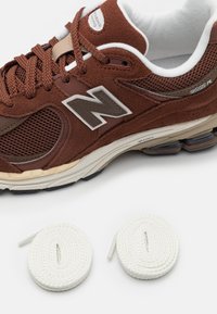 Närbild av en brun New Balance 2002R-sneaker med texturerad mocka, nätpaneler och två vridna vita skosnören på en vit bakgrund.