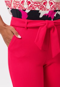 Pink textured pants with a tie belt at the waist and side pockets. The fabric features a patterned texture, enhancing the garment's visual depth.