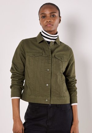 Femme portant une veste boutonnée vert olive sur un col roulé rayé noir et blanc, debout devant un fond clair uni.