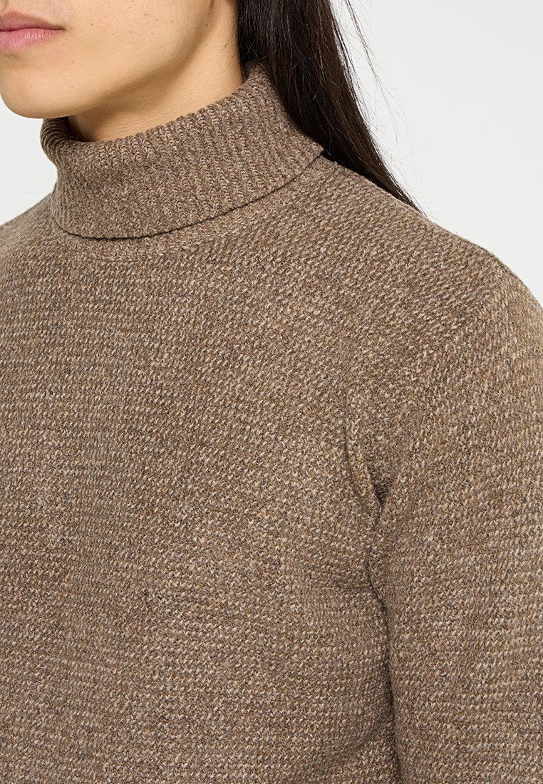 Donna che indossa un maglione a collo alto in maglia di colore marrone, mostrando il lato del collo, della spalla e parte del suo viso con lunghi capelli scuri.