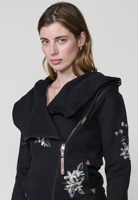 Sudadera con capucha de cremallera negra con textura de espiga, bordado floral en los hombros y detalles de cuero en la cremallera y los bolsillos.