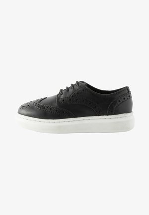 Chaussure en cuir noire avec des détails en brogue, bout rond et semelle épaisse blanche. Dispose de six œillets pour les lacets et d'accents texturés sur la tige.